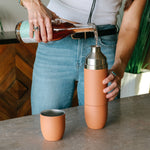 A person pours amber liquid from a glass bottle with a light green topographical label into a salmon-colored Parkit travel tumbler and matching cup on a grey marbled counter