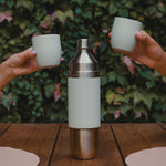 Photography of a two-toned stainless steel and matte sage Parkside Flask bottle on a wooden table, with two hands holding matching sage cups raised on either side, against a lush green ivy wall