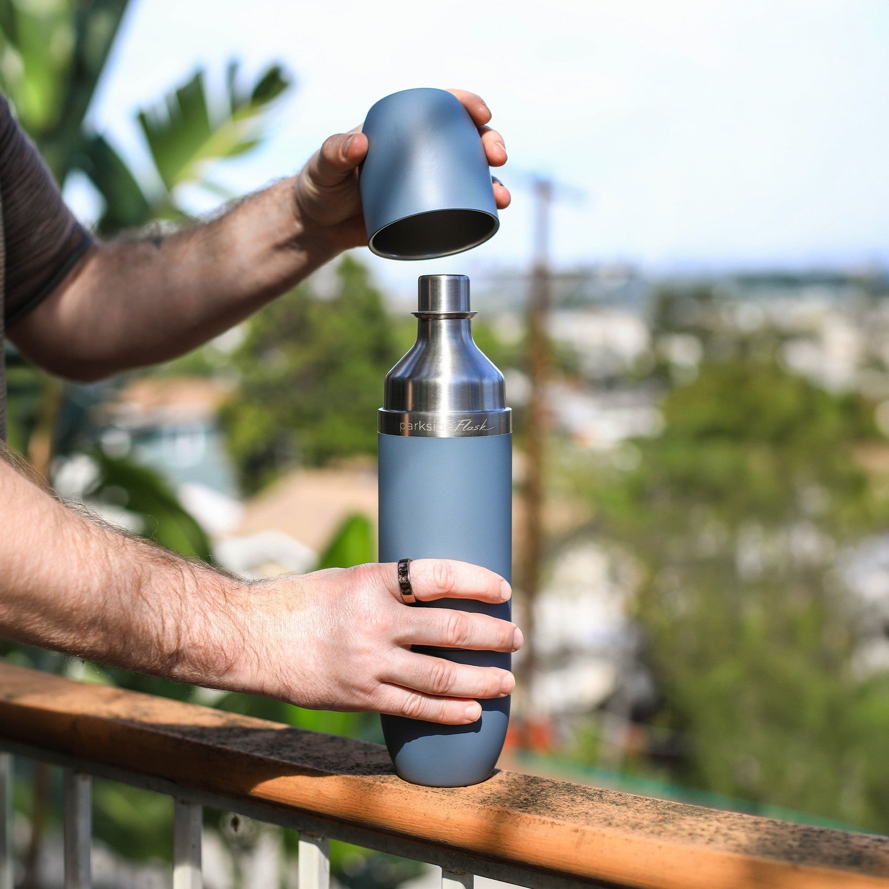 Photograph of hands opening a blue-gray Parkside Flask water bottle with a shining stainless steel top, a textured ring visible on a finger, resting on a wooden balcony railing overlooking a blurred green city landscape