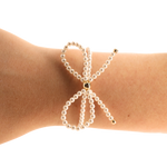 Photograph of a delicate pearl bow bracelet featuring a central gold bead and small gold tips on a person's wrist against a stark black background