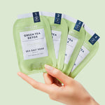 Product photography of a hand holding four light green Voesh New York Pedi in a Box Green Tea Detox 4-step packets prominently featuring "Vegan Clean" labels and details like Sea Salt Soak and Organic Olive Oil and Green Tea Extract
