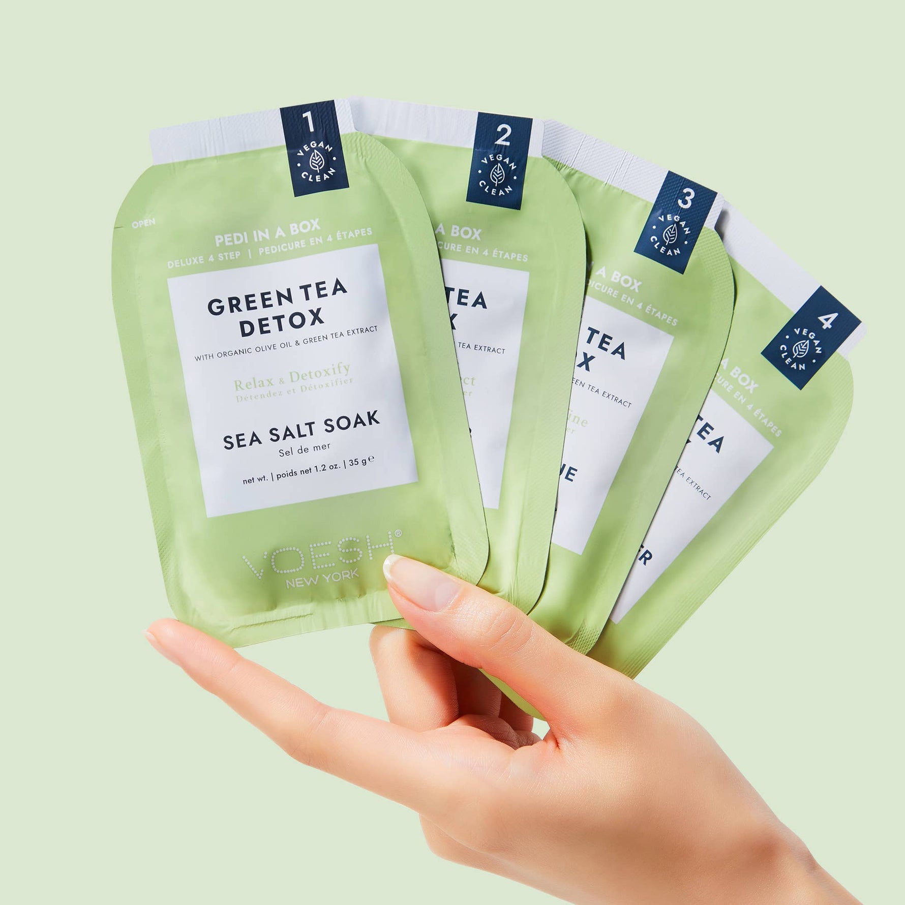 Product photography of a hand holding four light green Voesh New York Pedi in a Box Green Tea Detox 4-step packets prominently featuring "Vegan Clean" labels and details like Sea Salt Soak and Organic Olive Oil and Green Tea Extract