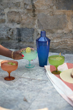 Lifestyle image showing a dark-skinned hand with a gold bracelet holding a pale green margarita with a lime slice, alongside a cobalt blue shaker, an orange jalapeño cocktail, and a bright green flamingo-stirred drink on a sunlit stone patio