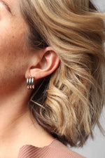 Close-up of a woman's ear with a shiny silver triple hoop earring, wavy blonde hair, and a blush pink ribbed top