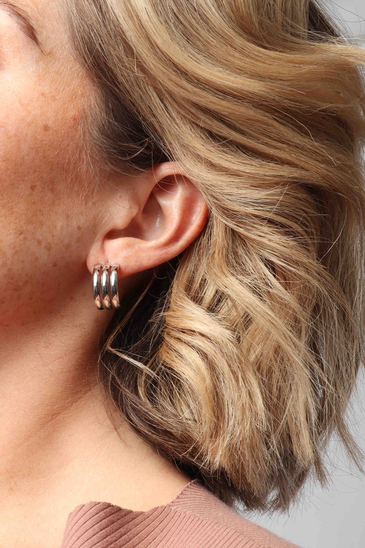 Close-up of a woman's ear with a shiny silver triple hoop earring, wavy blonde hair, and a blush pink ribbed top