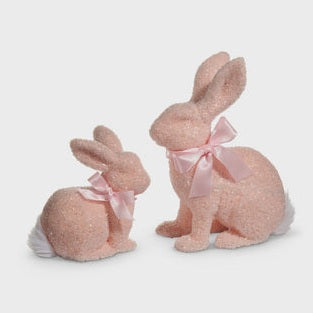 Two decorative pink glitter bunnies, one larger and sitting upright with a pink satin bow, the other smaller and crouching with a matching bow, both featuring white fluffy tails