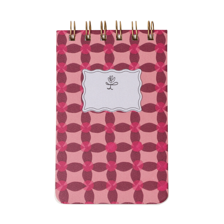 A small spiral-bound notebook with a pink and dark pink geometric quatrefoil pattern cover, a white scalloped label featuring a black floral icon, and gold coil binding
