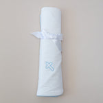 White rolled baby blanket tied with a white satin ribbon featuring a light blue embroidered cross and scallop stitching on a neutral background
