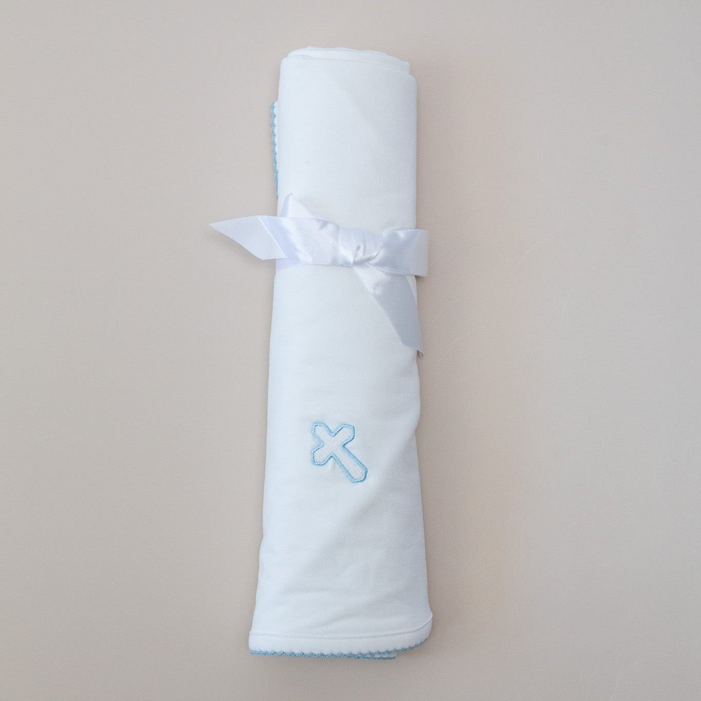 White rolled baby blanket tied with a white satin ribbon featuring a light blue embroidered cross and scallop stitching on a neutral background