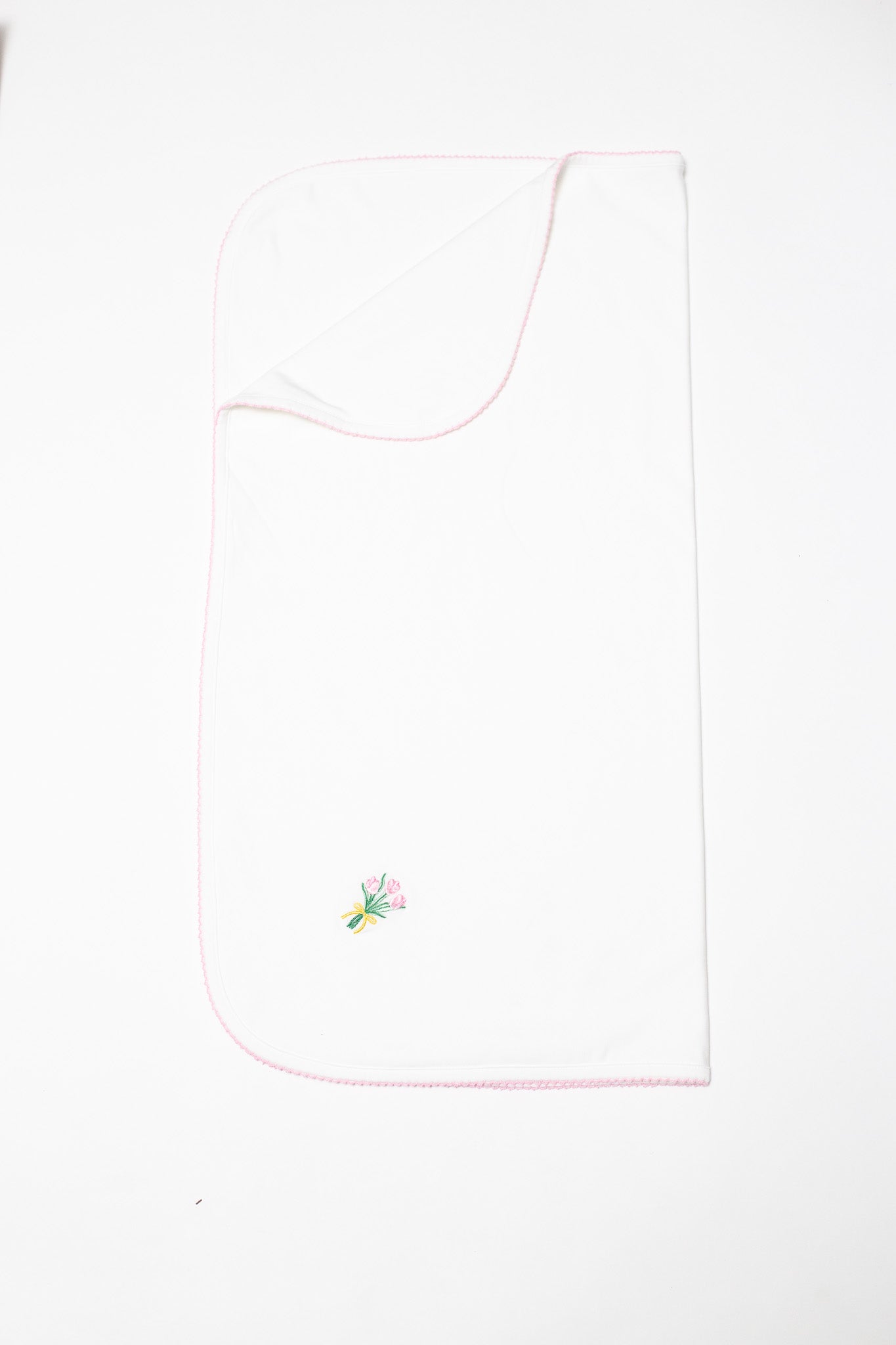 White baby blanket on a plain background with a delicate pink scalloped edge and a small embroidered bouquet of pink tulips tied with a yellow ribbon