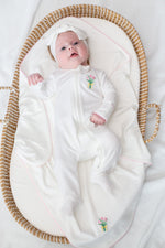 A newborn baby in a white footed sleeper with pink tulip embroidery and a matching white bow headwrap lies in a woven Moses basket lined with a white blanket trimmed in pink and featuring another pink tulip design