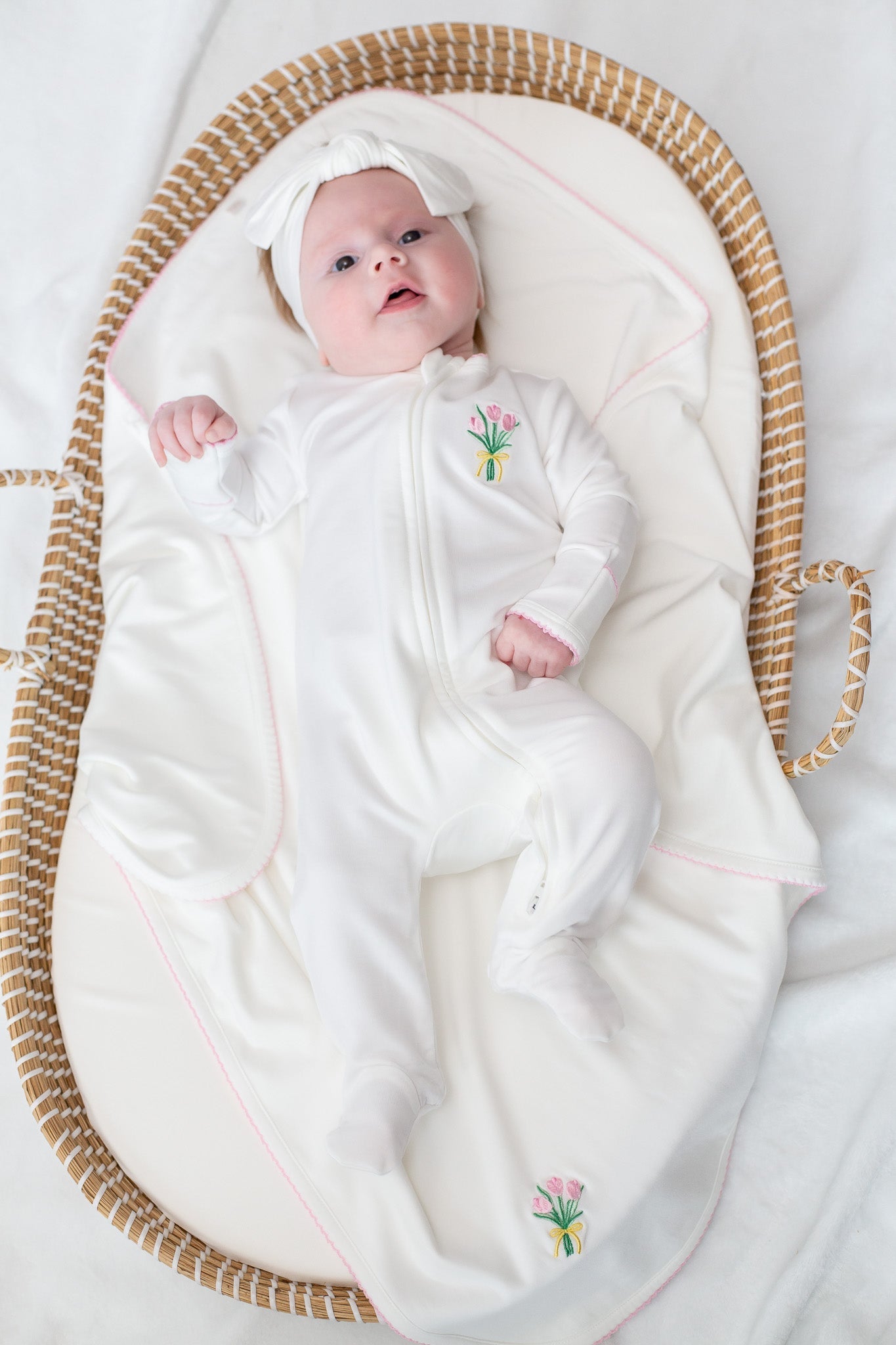 A newborn baby in a white footed sleeper with pink tulip embroidery and a matching white bow headwrap lies in a woven Moses basket lined with a white blanket trimmed in pink and featuring another pink tulip design