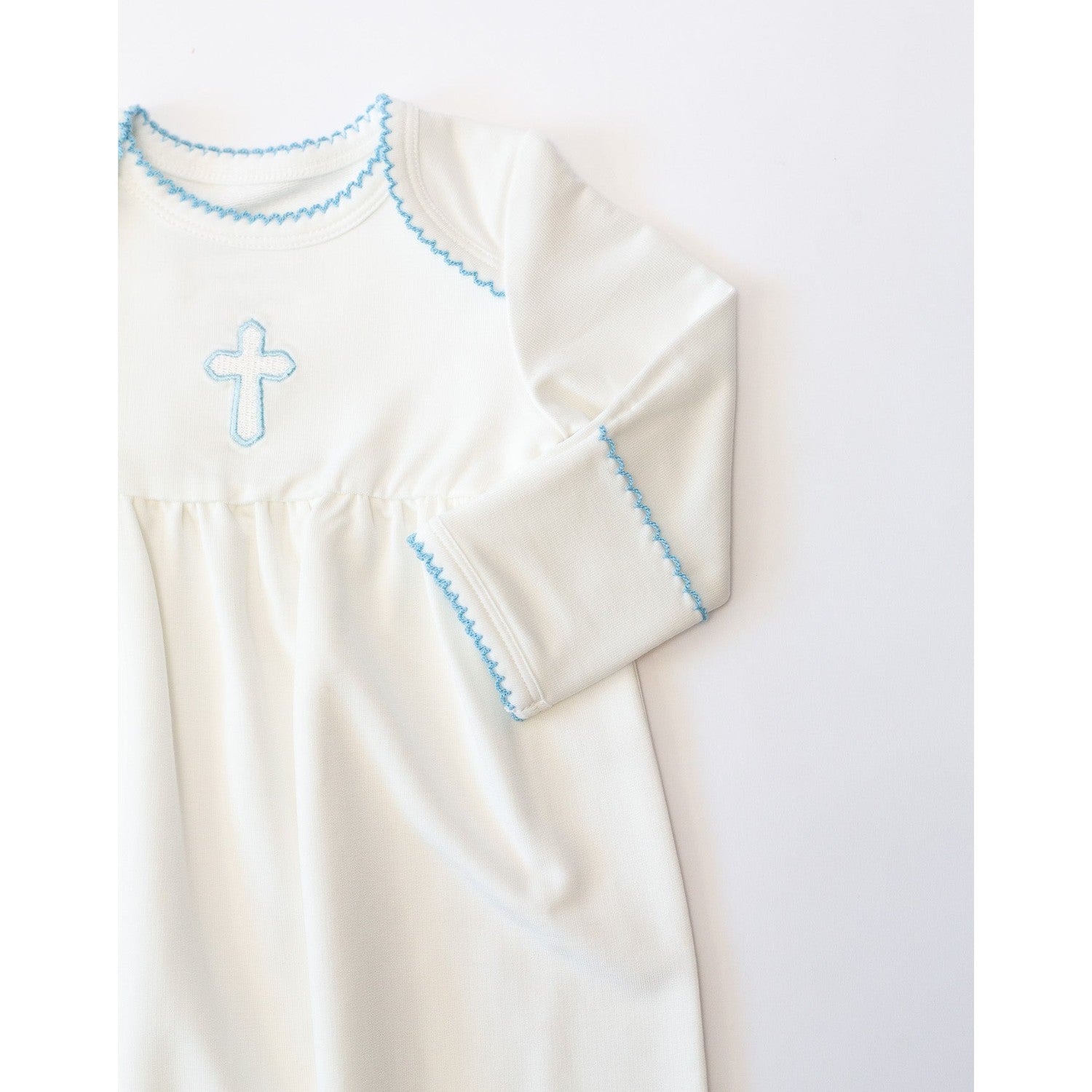 Studio photograph of a white cotton baby gown featuring a light blue embroidered cross and delicate scalloped stitching along the neckline and sleeve cuffs set against a minimalist white background