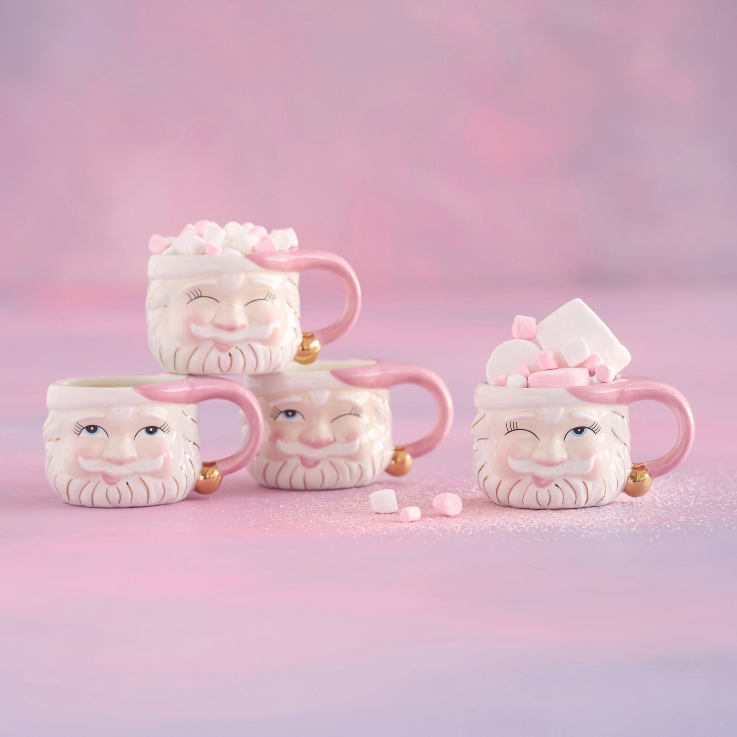 Four Santa Claus shaped mugs with pink accents filled with marshmallows on a pink background