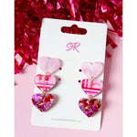 Acrylic earrings by Sandy Rizzo, a pair of tiered heart dangle earrings on a white display card set against a vibrant pink background with shimmering red tinsel, featuring alternating light pink, plaid patterned, and chunky glitter-filled hearts