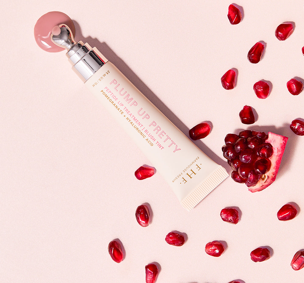 Farmhouse Fresh Plump Up Pretty Peptide Lip Treatment Blush Tint tube with a heart-shaped applicator dispensing a pink dollop, alongside vibrant red pomegranate seeds and a cut pomegranate slice on a light pink background