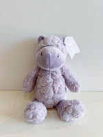 Cuddle + Kind lavender purple fluffy Dolly the Hippo plush toy with embroidered eyes and nose sits on a light surface against a white wall showing its brand tag