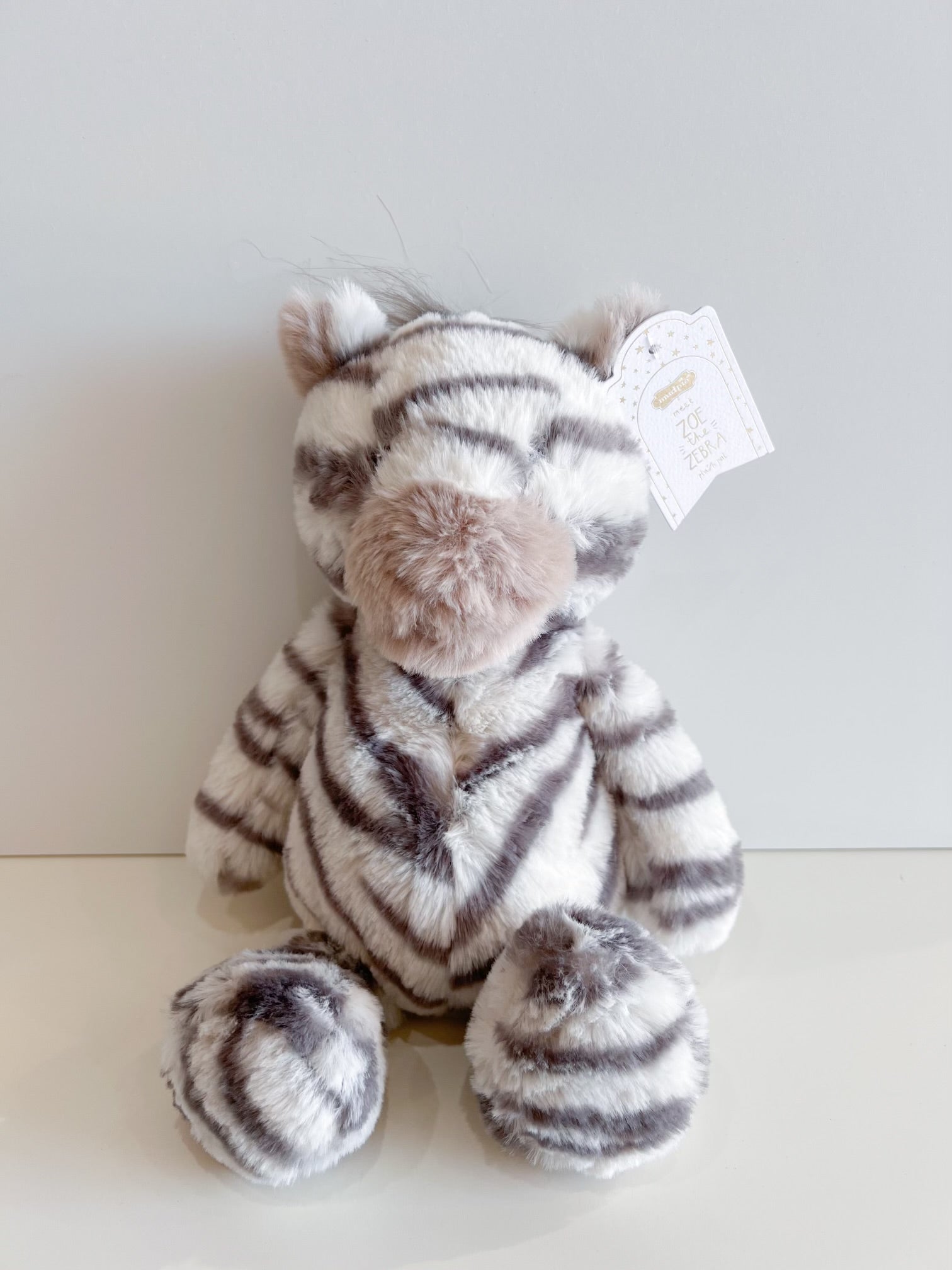Mud Pie plush toy Zoe the Zebra sitting on a light surface featuring soft white and gray striped fur, a fluffy pinkish-brown nose, and a brand tag