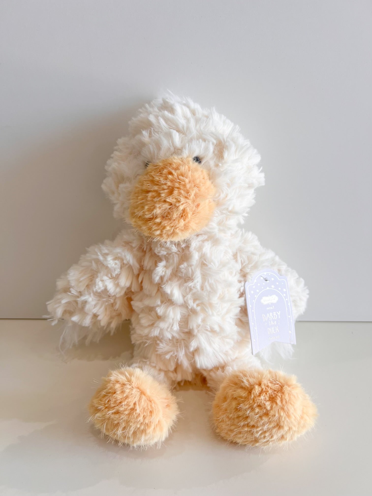 Plush toy of a shaggy white duck with an orange bill and feet seated against a plain white background featuring a light purple tag that reads 'meet DARBY the DUCK'