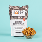 Product photography of a white and brown patterned Poppy Hand-Crafted Popcorn bag with an orange poppy logo and "Cinnamon Bourbon Pecan" flavor text, standing next to a clear bowl filled with golden caramelized popcorn and visible whole pecans on a bright light blue background