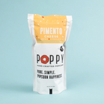 Studio shot of a white stand-up pouch of Poppy Hand-Crafted Popcorn Pimento Cheese flavor made in Asheville NC, featuring a red poppy logo and yellow top against a light blue background