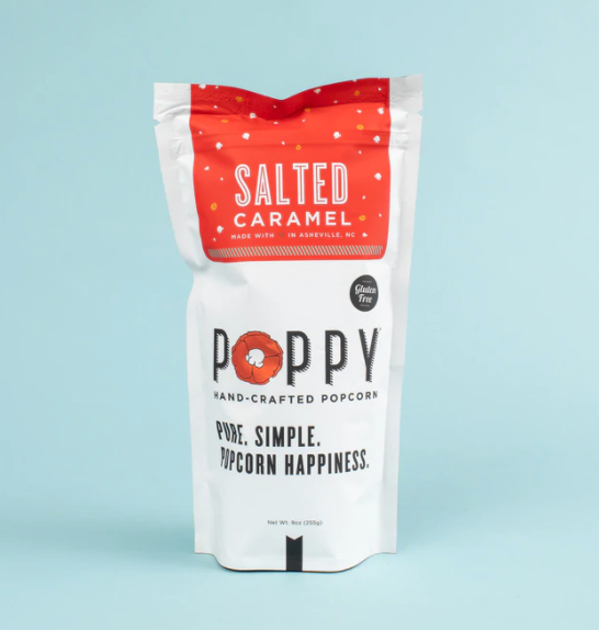 Product photography of a white stand-up pouch of Poppy Salted Caramel Hand-Crafted Popcorn made in Asheville NC featuring a vibrant red top label with white speckles an orange poppy flower in the logo and a gluten-free badge against a solid light blue background