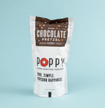 Product photography of a white Poppy Hand-Crafted Popcorn Dark Chocolate Pretzel pouch featuring a prominent red poppy logo and dark brown top label with scattered pretzel shapes, indicating it's made in Asheville NC with craft chocolate from French Broad Chocolate, all against a soft blue background