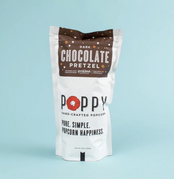 Product photography of a white Poppy Hand-Crafted Popcorn Dark Chocolate Pretzel pouch featuring a prominent red poppy logo and dark brown top label with scattered pretzel shapes, indicating it's made in Asheville NC with craft chocolate from French Broad Chocolate, all against a soft blue background