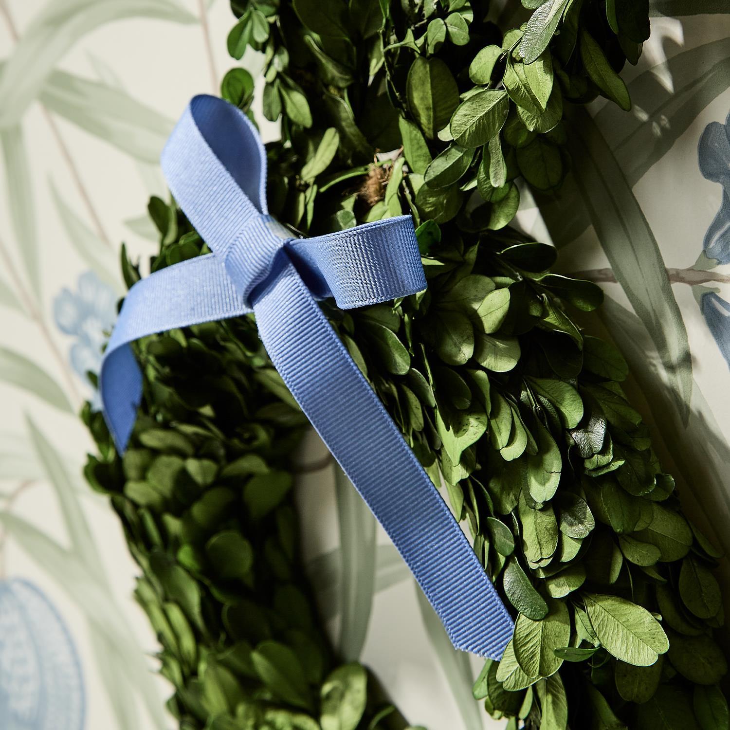 A preserved boxwood wreath adorned with a light blue grosgrain ribbon bow set against a soft floral wallpaper featuring subtle blue and green botanical patterns