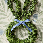 Green boxwood bunny wreath with a light blue grosgrain ribbon bow hanging on a patterned wallpaper featuring green leaves and delicate blue flowers