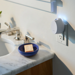 Photograph of a white Pura smart fragrance diffuser glowing blue plugged into a wall outlet in a modern bathroom, positioned next to a white marble vanity with a chrome faucet and an indigo blue soap dish holding a light brown bar of soap