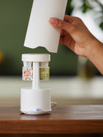 A hand places a ribbed white cover onto a sleek white fragrance diffuser on a wooden table, featuring two colorful Anthropologie refills: White Amber Mandarin with an orange design and Honeycrisp Oakwood with green leaves, illuminated by a subtle blue light