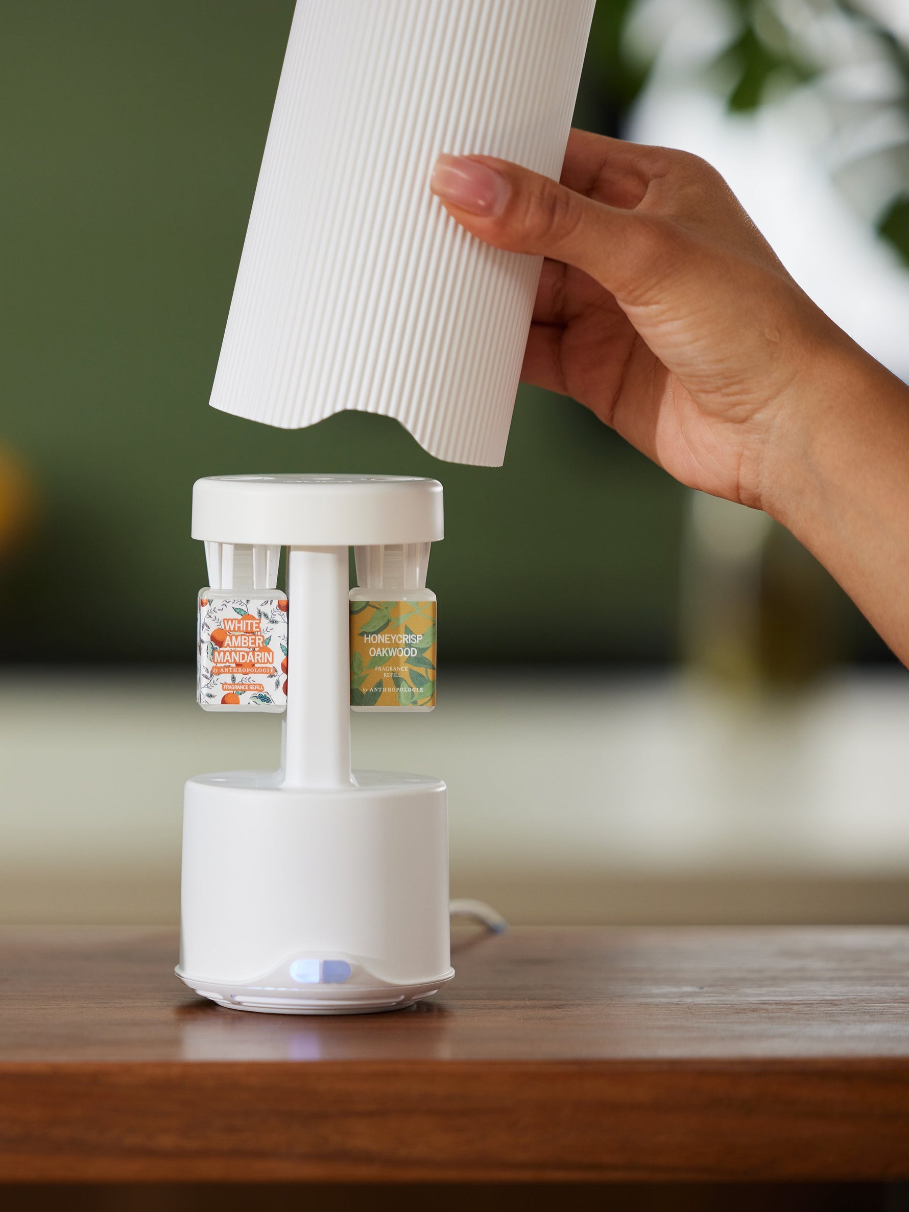 A hand places a ribbed white cover onto a sleek white fragrance diffuser on a wooden table, featuring two colorful Anthropologie refills: White Amber Mandarin with an orange design and Honeycrisp Oakwood with green leaves, illuminated by a subtle blue light