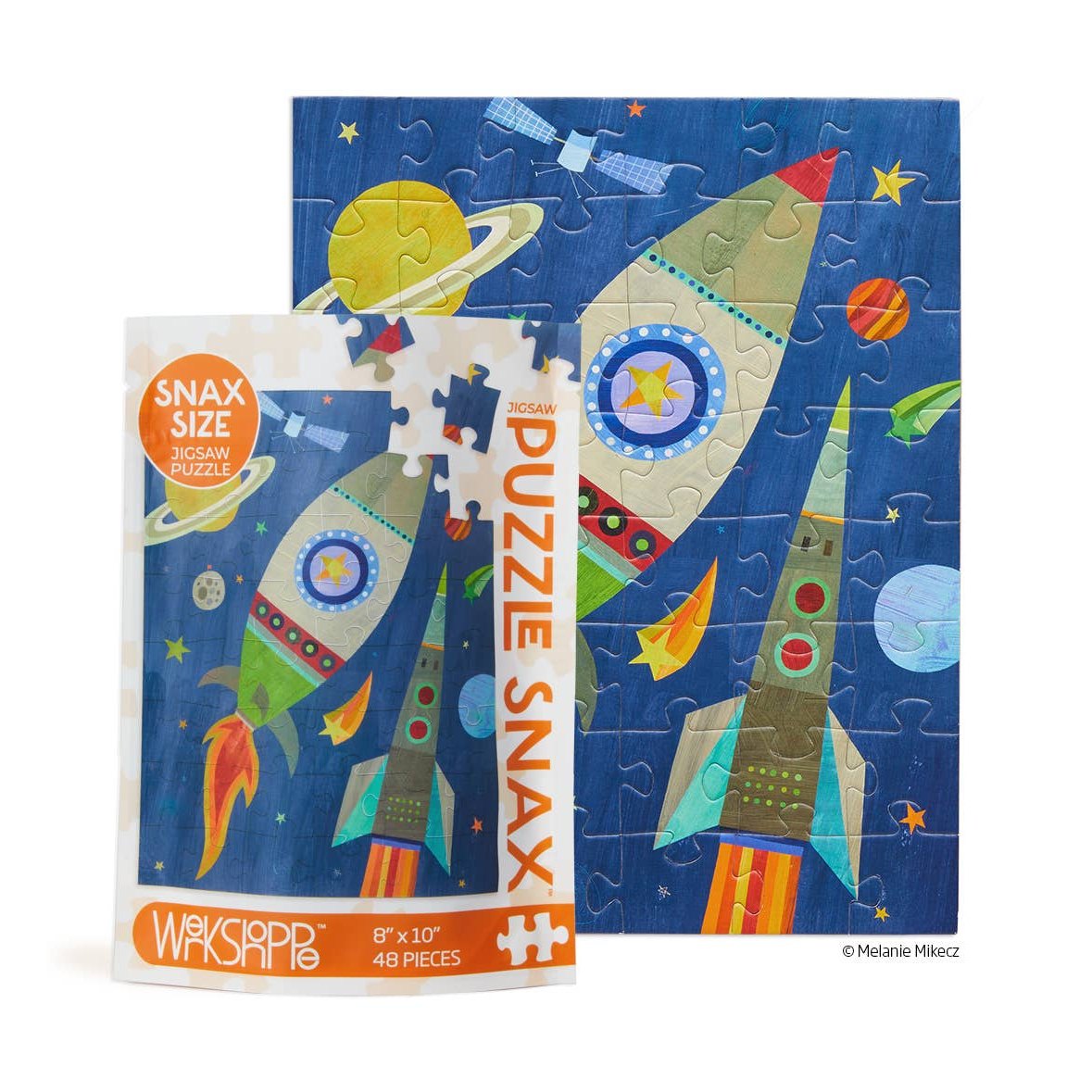 Jigsaw puzzle by Melanie Mikecz depicting a deep blue outer space scene featuring a green and brown rocket with orange exhaust flames, a gray and teal rocket with red portholes, and iconic Saturn with its rings, displayed alongside its "Snax Size" packaging