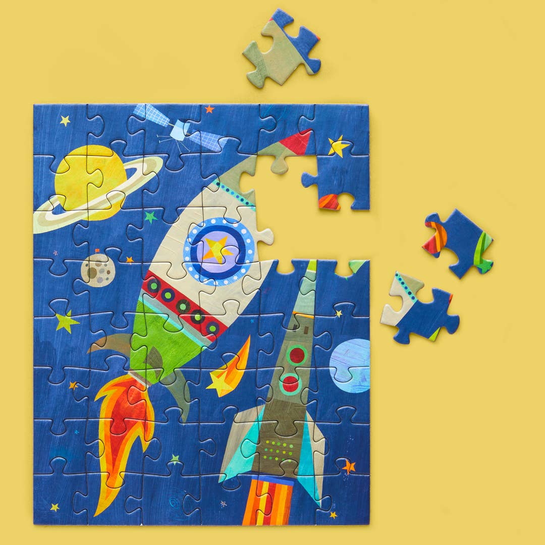 Jigsaw puzzle depicting a partially completed cosmic scene in outer space with a large white and green rocket launching towards a yellow Saturn with rings and a small grey moon, alongside a second stylized rocket and several scattered loose pieces on a yellow background