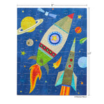 Jigsaw puzzle by Melanie Mikecz depicting a colorful outer space scene featuring two stylized rockets a yellow Saturn with rings and a small satellite against a deep blue starry background with one rocket having fiery orange exhaust and the other striped with red and orange