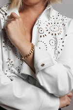 Photograph showing a person's arm and chest in a white shirt featuring intricate laser-cut patterns, a shiny gold Cuban link bracelet, and silver cuff snap buttons