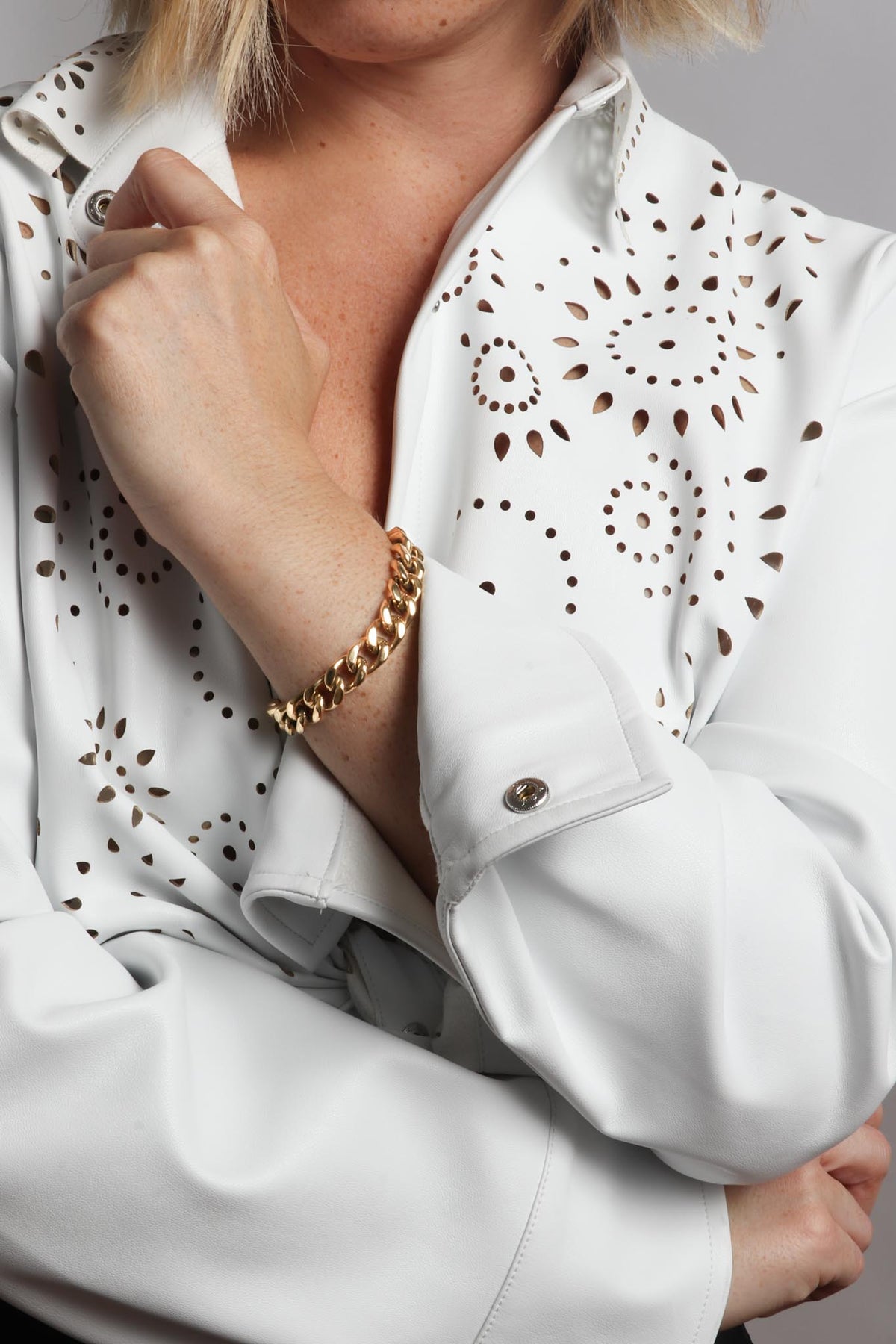 Photograph showing a person's arm and chest in a white shirt featuring intricate laser-cut patterns, a shiny gold Cuban link bracelet, and silver cuff snap buttons
