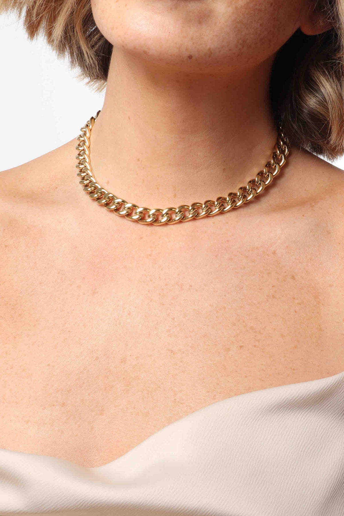 A close-up of a model's neck featuring a chunky gold curb chain necklace on freckled skin over a cream top