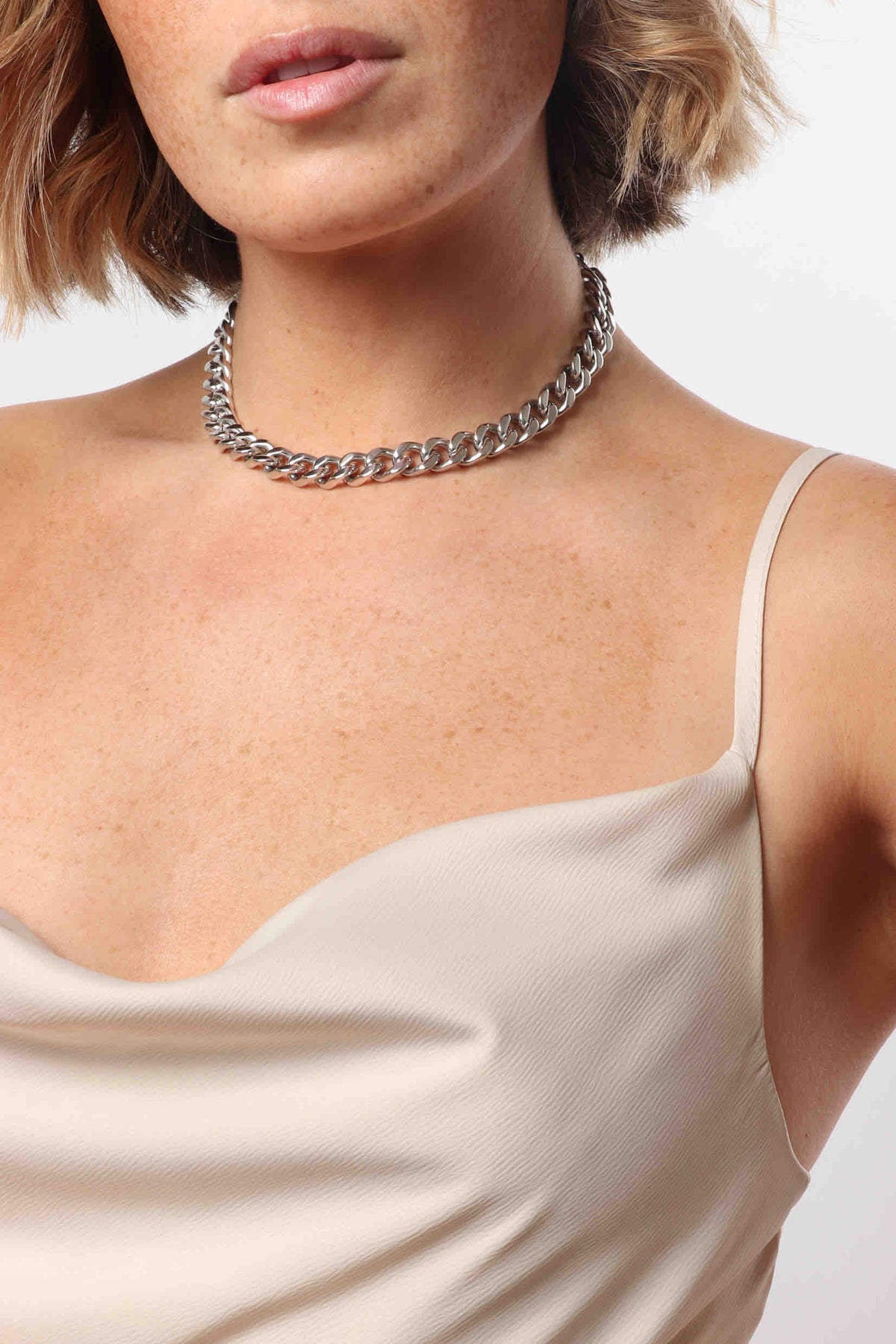 Close-up of a woman's upper body featuring a gleaming silver cuban link choker necklace and a champagne silk cowl neck slip dress, revealing freckled skin and blonde hair