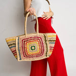 A person in red wide-leg pants and a white crop top holding a large handmade straw tote bag featuring a vibrant multi-colored granny square crochet pattern with a prominent green center