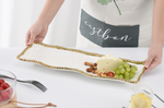 A person in a white apron presents a white rectangular serving platter with a distinctive gold beaded wavy edge, featuring a wedge of Swiss cheese, green grapes, and mixed nuts, with cherry tomatoes and lemon water on the side