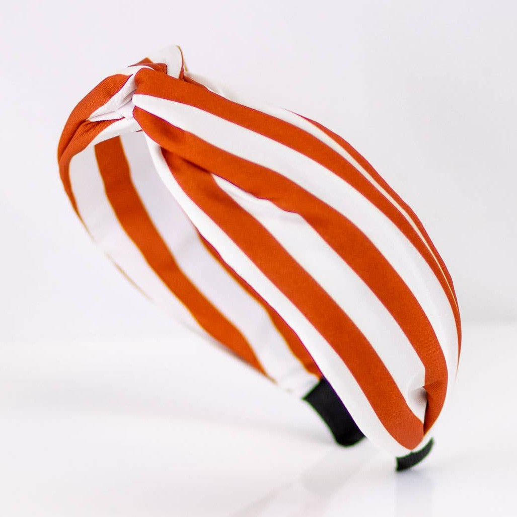 Photograph of a knotted fabric headband against a white background featuring vibrant burnt orange and white diagonal stripes with a central top twist and black ends