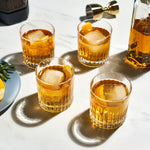 Studio photography of four amber whiskey cocktails in fluted glasses with large square ice and orange peels on a white marble surface, beside a gold jigger and fresh rosemary