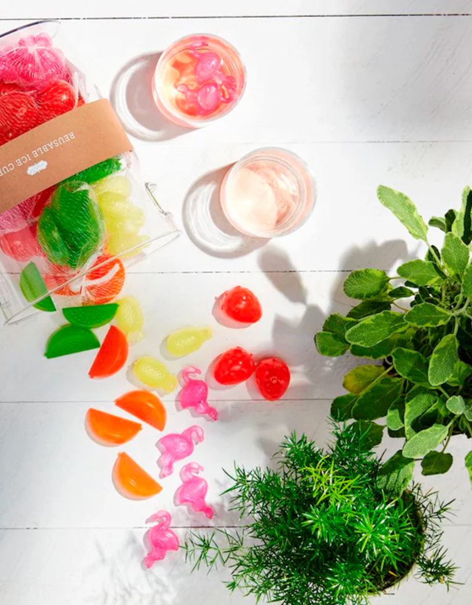Reusable plastic ice cubes shaped like fruit and flamingos spilled on a white table next to two glasses and plants
