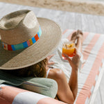 A woman wearing a wide-brimmed straw hat with a vibrant multi-colored striped band and sun emblem relaxes on a peach and white striped lounge chair, holding a refreshing orange cocktail on a sunny beach or poolside deck