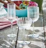 Photograph of three clear ribbed drinking glasses, a flute, coupe, and stemmed glass, on a weathered wooden outdoor table with a vibrant turquoise floral bowl holding brown wafer rolls and a pink napkin