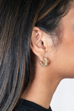 Close-up of a woman's ear framed by dark brown highlighted hair and a black sweater, featuring a small gold twisted hoop earring embedded with sparkling clear crystals