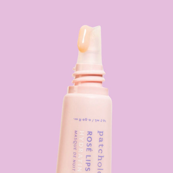Product photography of a pale pink Patchology Rosé Lips Hydrating Masque de Nuit tube with a clear angled applicator displaying a shiny peachy-pink product bead against a solid lavender background