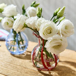 Two glass vases holding white Lisianthus flowers and green buds on a sunlit wooden table, one vase intricately striped with red and the other with blue